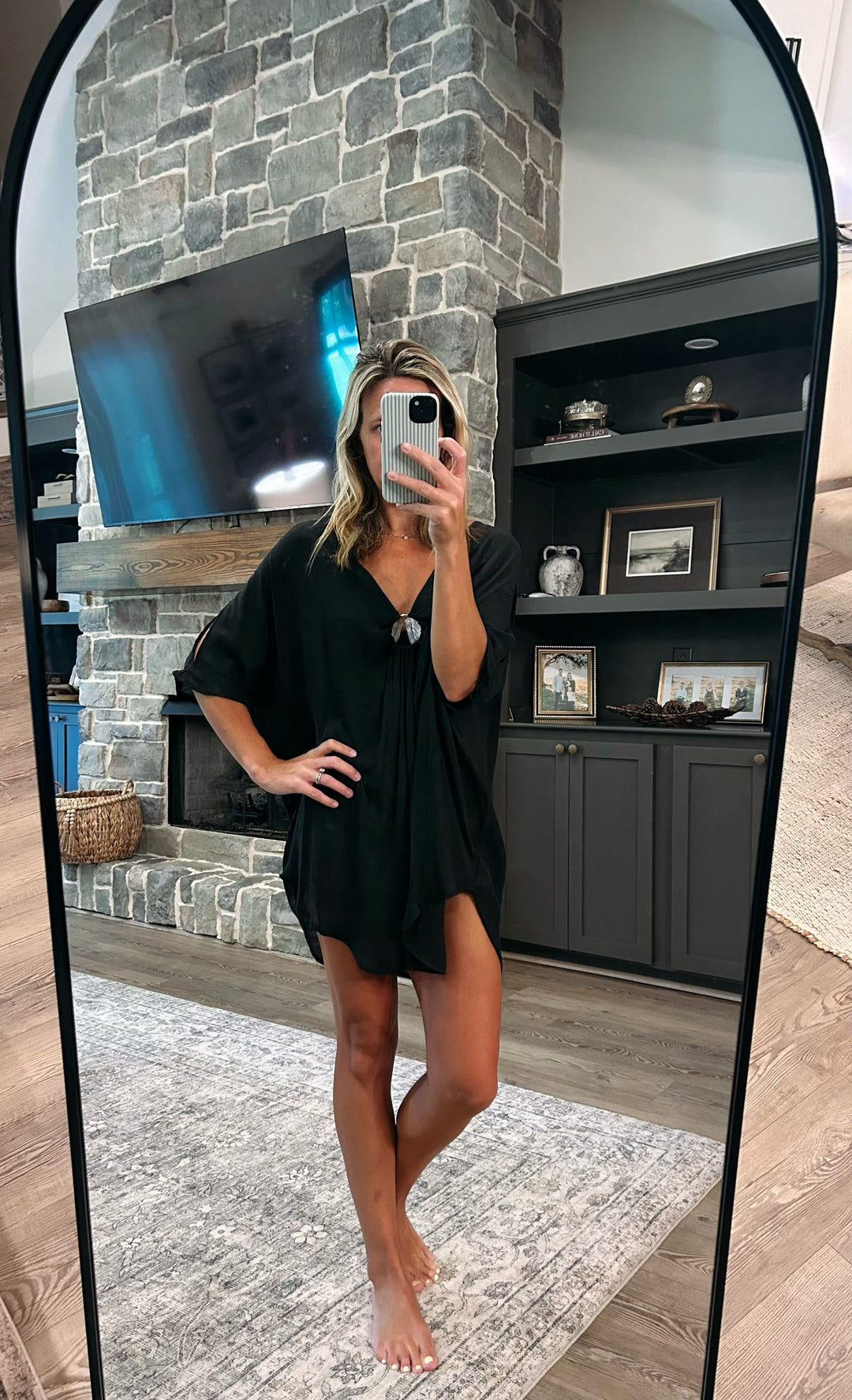 A woman takes a mirror selfie in a living room wearing a black short-sleeve V-neck tunic dress or cover-up. The background features a stone fireplace, a mounted TV, and a dark wood shelving unit.