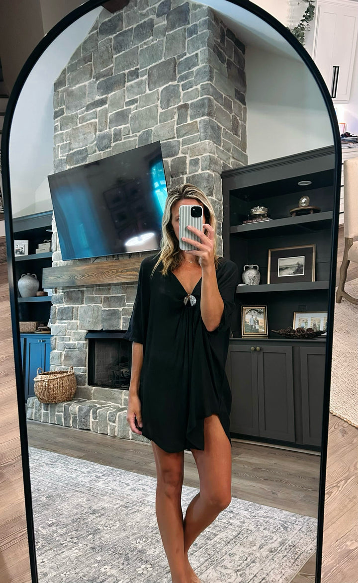 A woman takes a mirror selfie in a living room wearing a black short-sleeve mini dress with a V-neckline and a small silver pendant at the center. The background features a gray stone fireplace, a mounted TV, and dark gray shelving.
