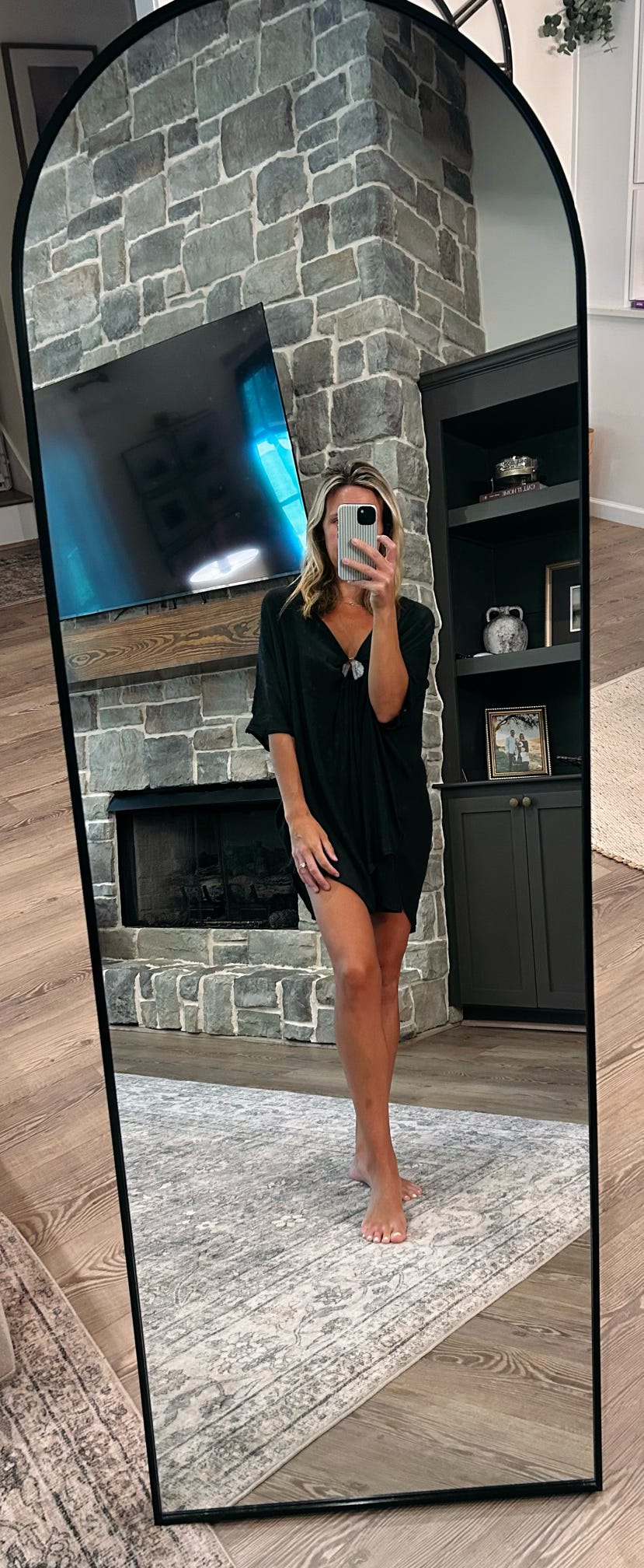 A woman takes a mirror selfie in a living room wearing a black short-sleeve mini dress. The dress has a loose, oversized fit and a small chest pocket.