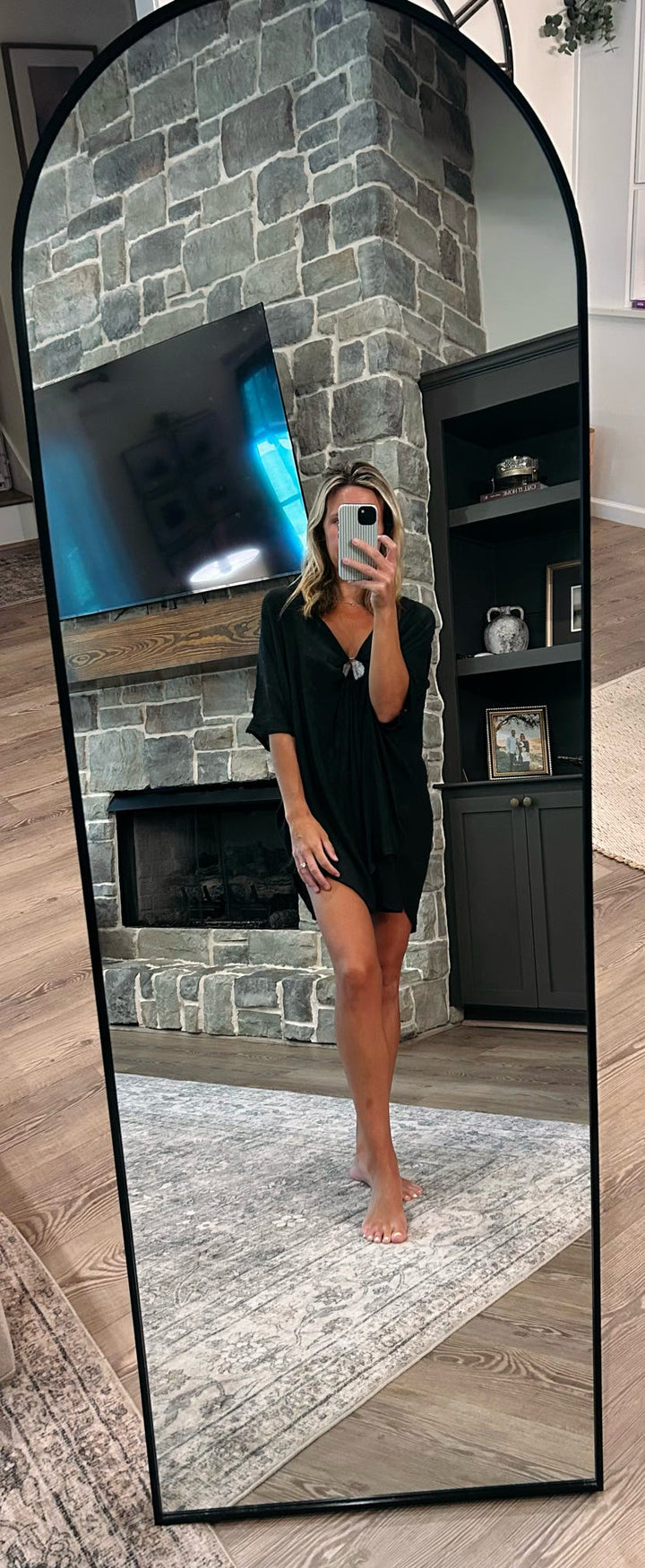 A woman takes a mirror selfie in a living room wearing a black short-sleeve mini dress. The dress has a loose, oversized fit and a small chest pocket.
