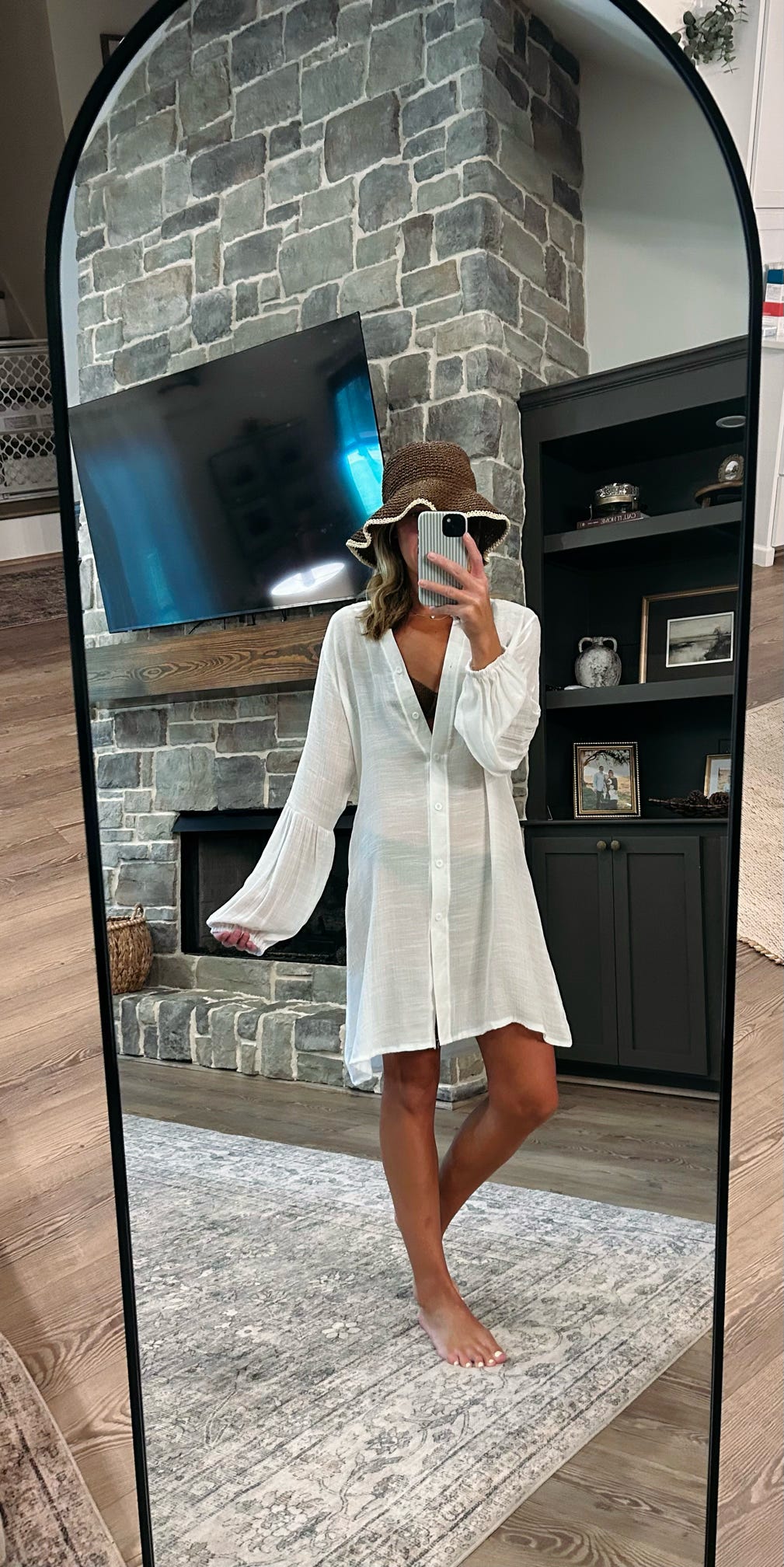 A woman takes a mirror selfie in a living room wearing a white long-sleeve button-up shirt dress with bell sleeves, a brown hat, and white sunglasses. The background features a gray stone fireplace, a black TV, and a dark wood shelving unit.