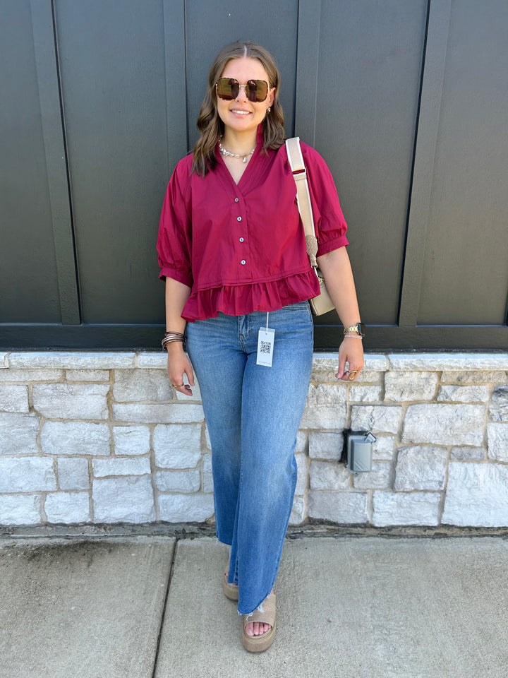 A woman wearing a burgundy ruffled short-sleeve blouse, blue jeans, and sunglasses stands outdoors against a dark gray paneled wall and stone base.
