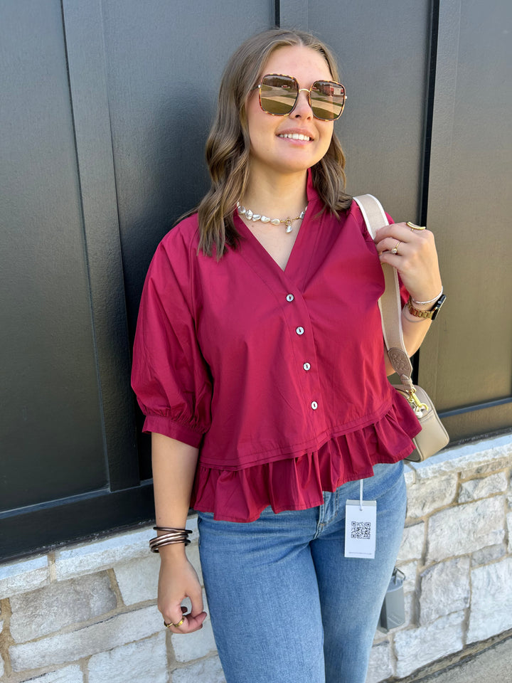 A woman wearing a burgundy puff-sleeve blouse with a ruffled hem, paired with blue jeans and sunglasses, standing against a dark gray paneled wall.