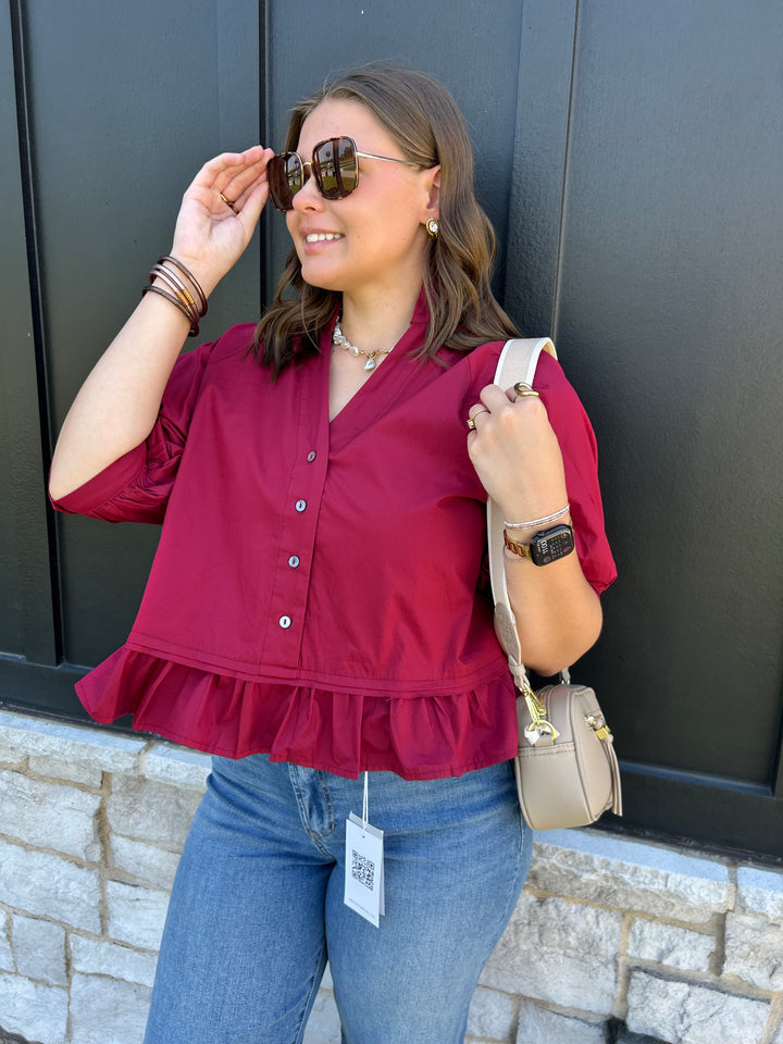 A woman wearing a burgundy short-sleeve button-up blouse with a ruffled hem, paired with blue jeans, sunglasses, and a beige shoulder bag, posing against a dark paneled wall.