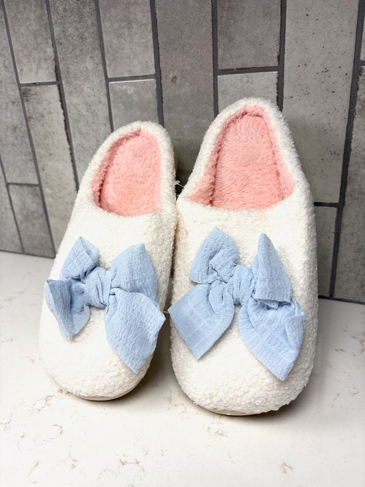 Cozy white fuzzy house slippers with soft pink lining and large light blue bow detail, placed on a countertop with gray tiled background.