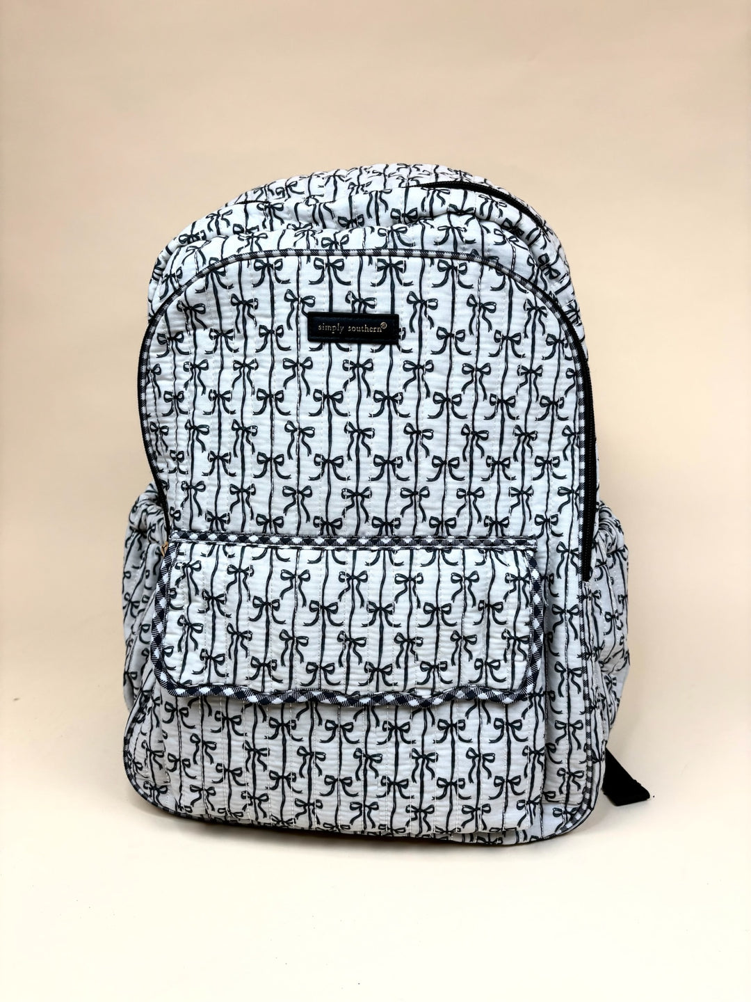 Simply Southern Black Bow Quilted Backpack featuring an allover black bow print, quilted construction, spacious zippered main compartment, front flap pocket, side pockets, and black trim on a cream studio background.