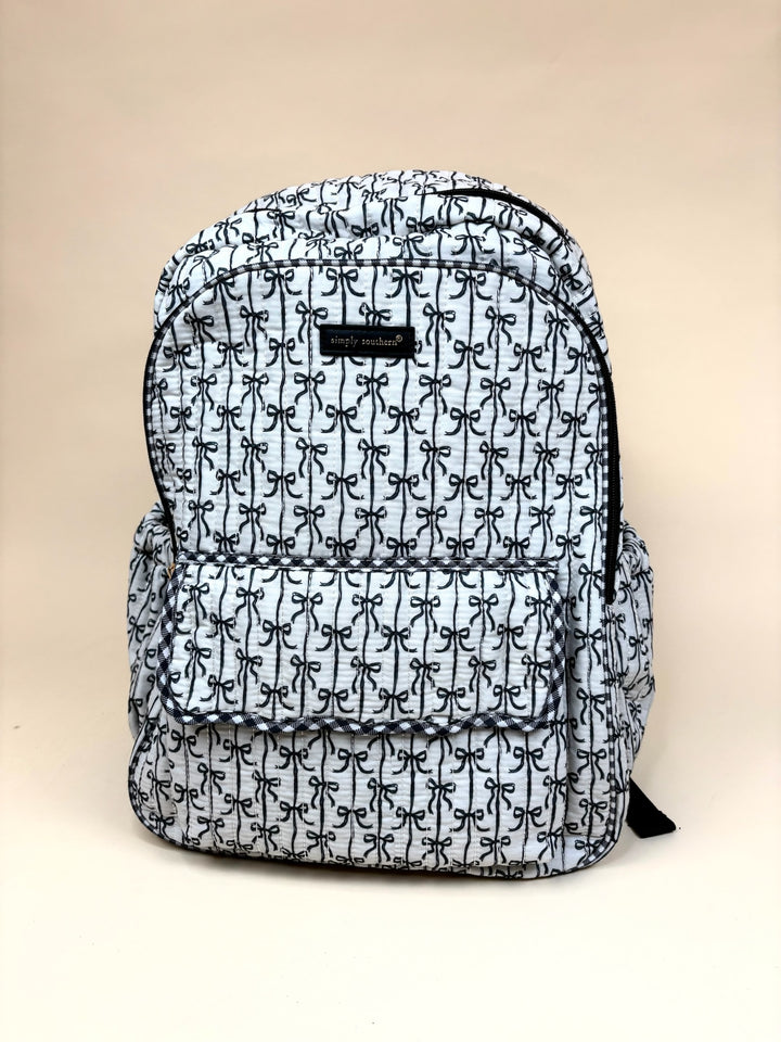 Simply Southern Black Bow Quilted Backpack featuring an allover black bow print, quilted construction, spacious zippered main compartment, front flap pocket, side pockets, and black trim on a cream studio background.