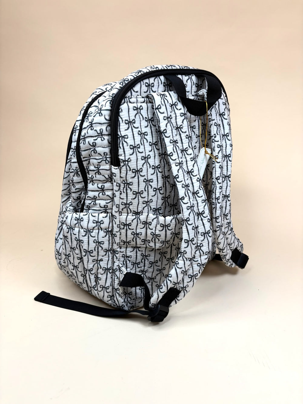 Back view of the Simply Southern Black Bow Quilted Backpack showing quilted bow print, padded adjustable shoulder straps, top carry handle, side pockets, and black trim on a cream studio background.
