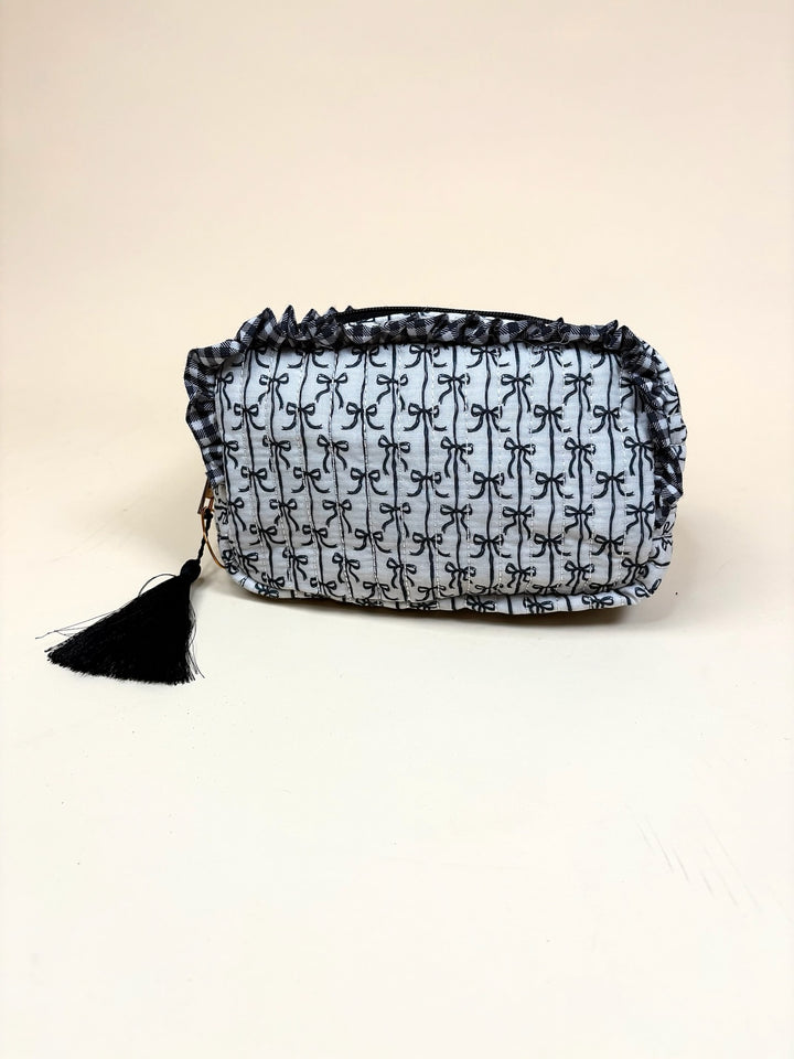 Simply Southern Black Bow Quilted Cosmo Bag featuring an allover black bow print, quilted design, ruffle trim, black tassel zipper pull, and zip-top closure on a cream studio background.
