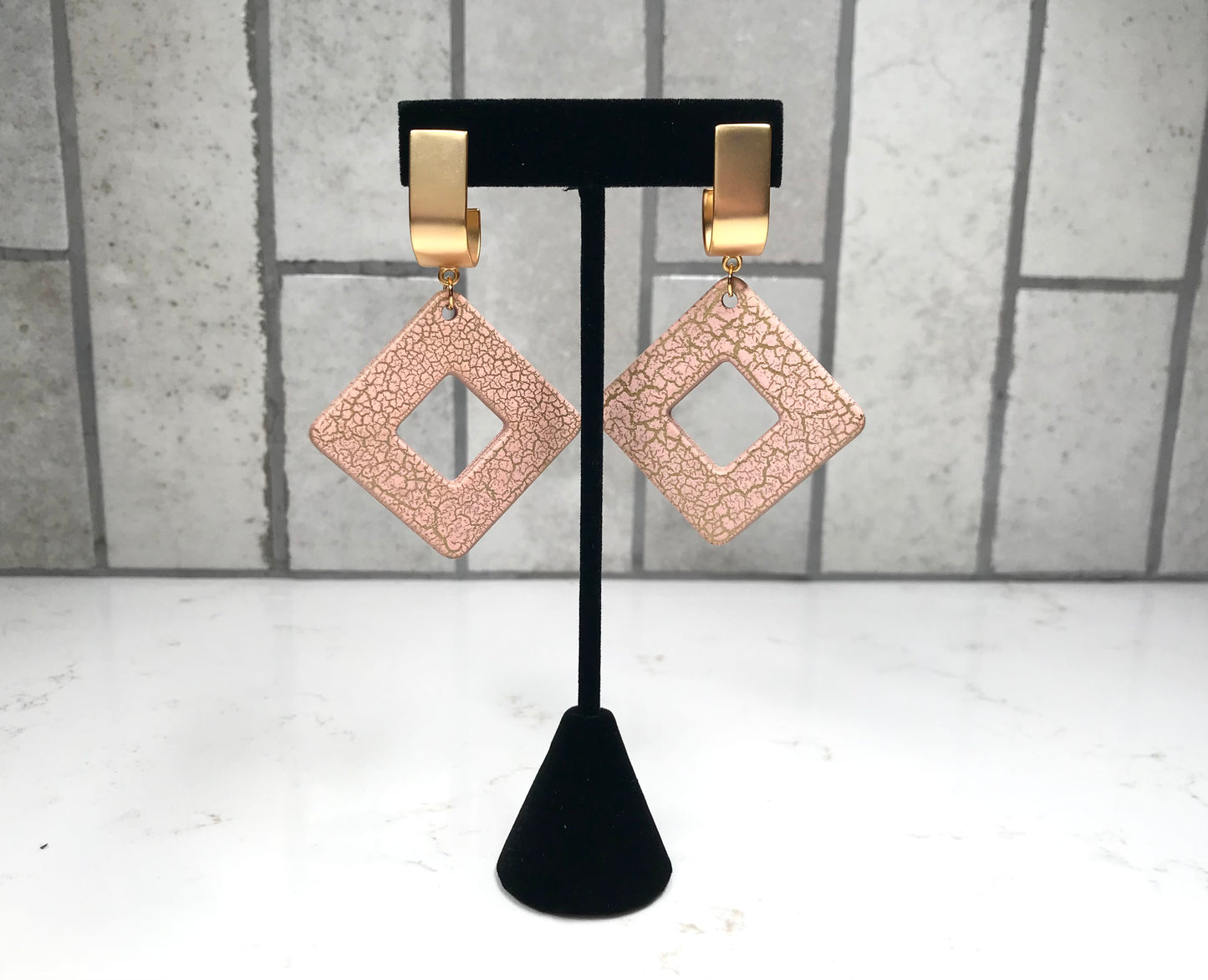 Rose gold square earrings on a black stand against a gray tiled wall.