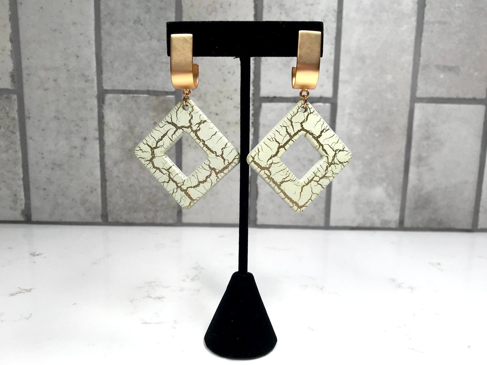 Pair of gold-framed diamond-shaped earrings on a black stand against a gray tiled wall.