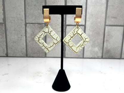Pair of gold-framed diamond-shaped earrings on a black stand against a gray tiled wall.