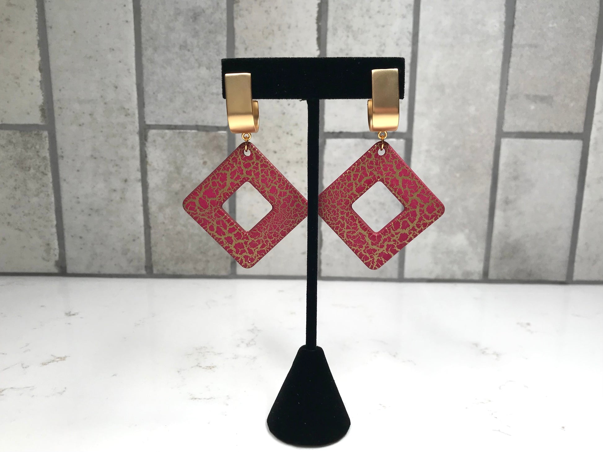 Red square earrings with gold accents on a black stand against a gray tiled wall.