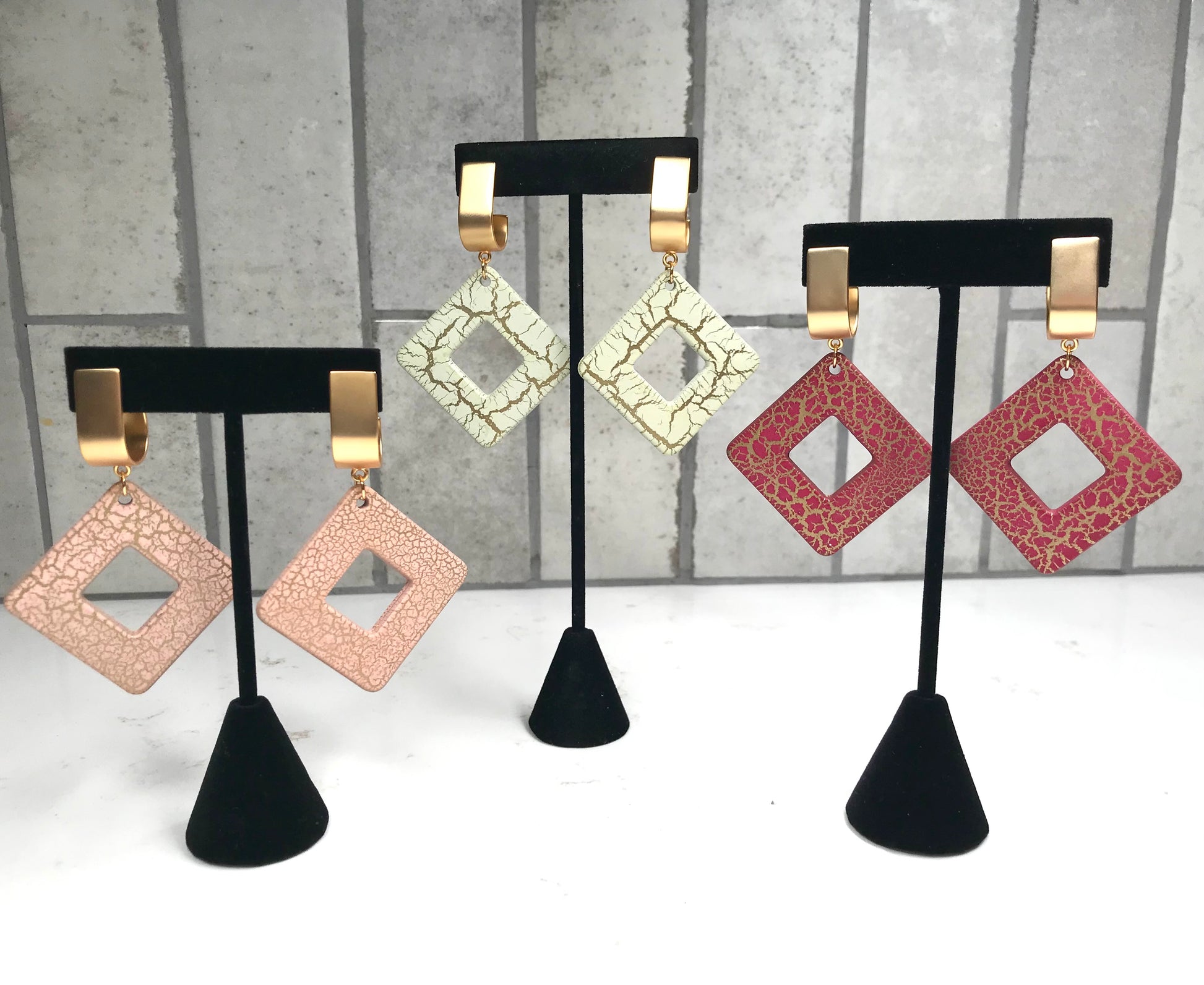 Set of square-shaped earrings on black stands against a gray tiled wall.