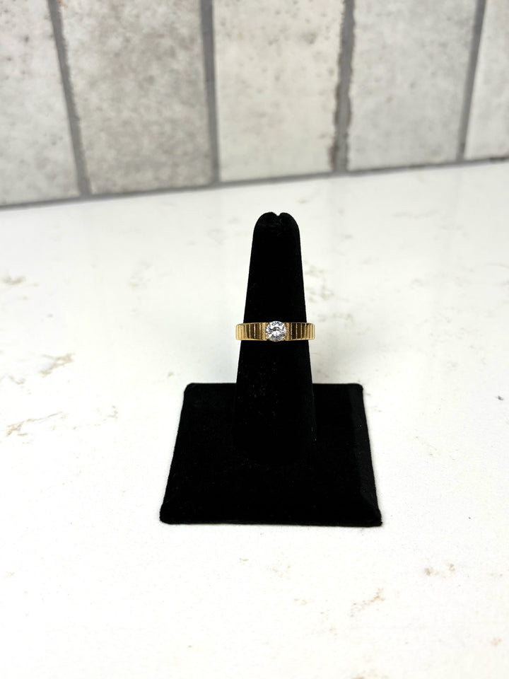 Gold ribbed band ring with a round clear crystal centerpiece displayed on a black velvet jewelry stand.
