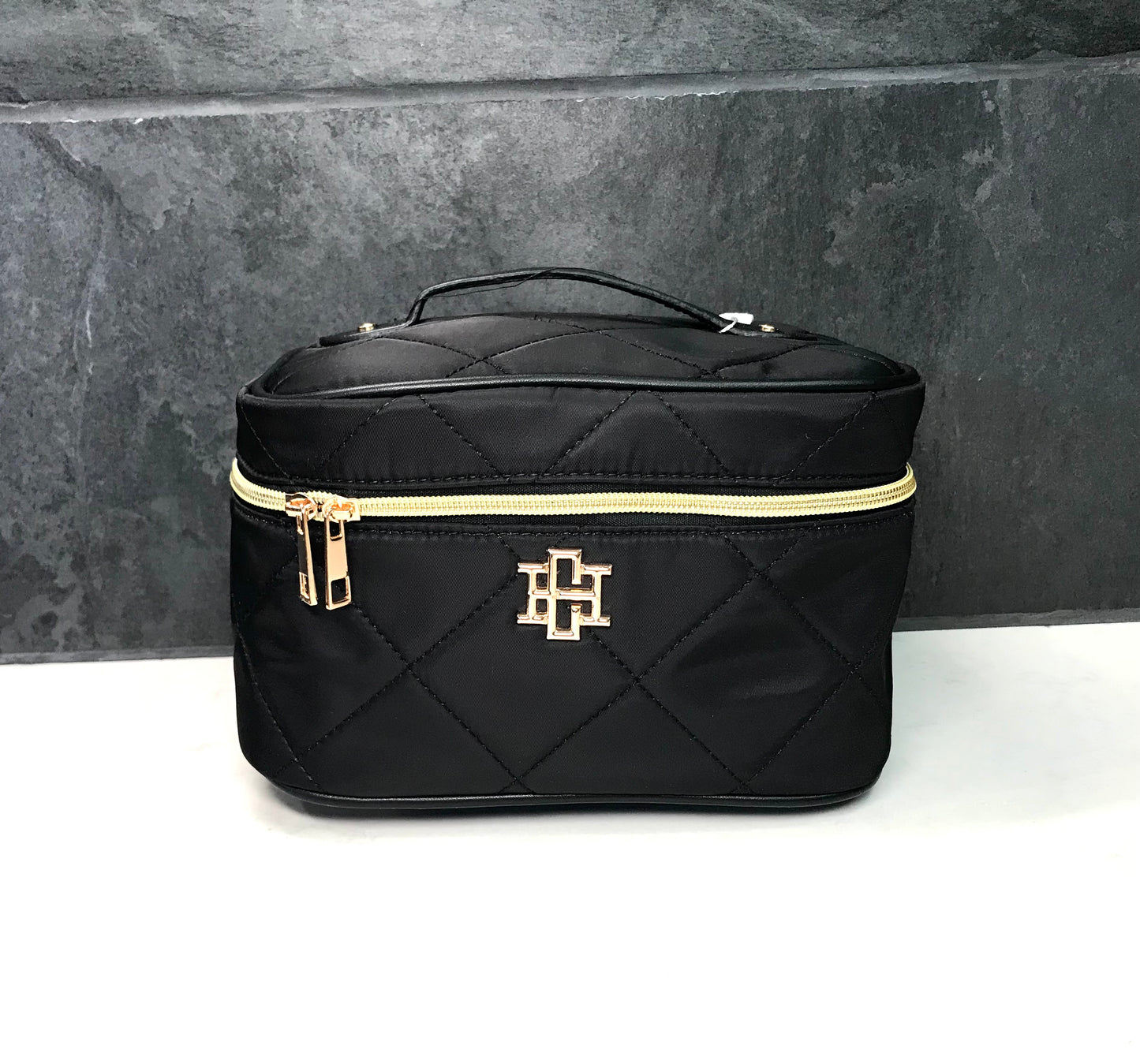 Black quilted makeup bag with gold zipper and logo on a gray background