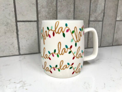 White mug with 'fa la la la' text and Christmas lights design on a marble surface with tiled wall background