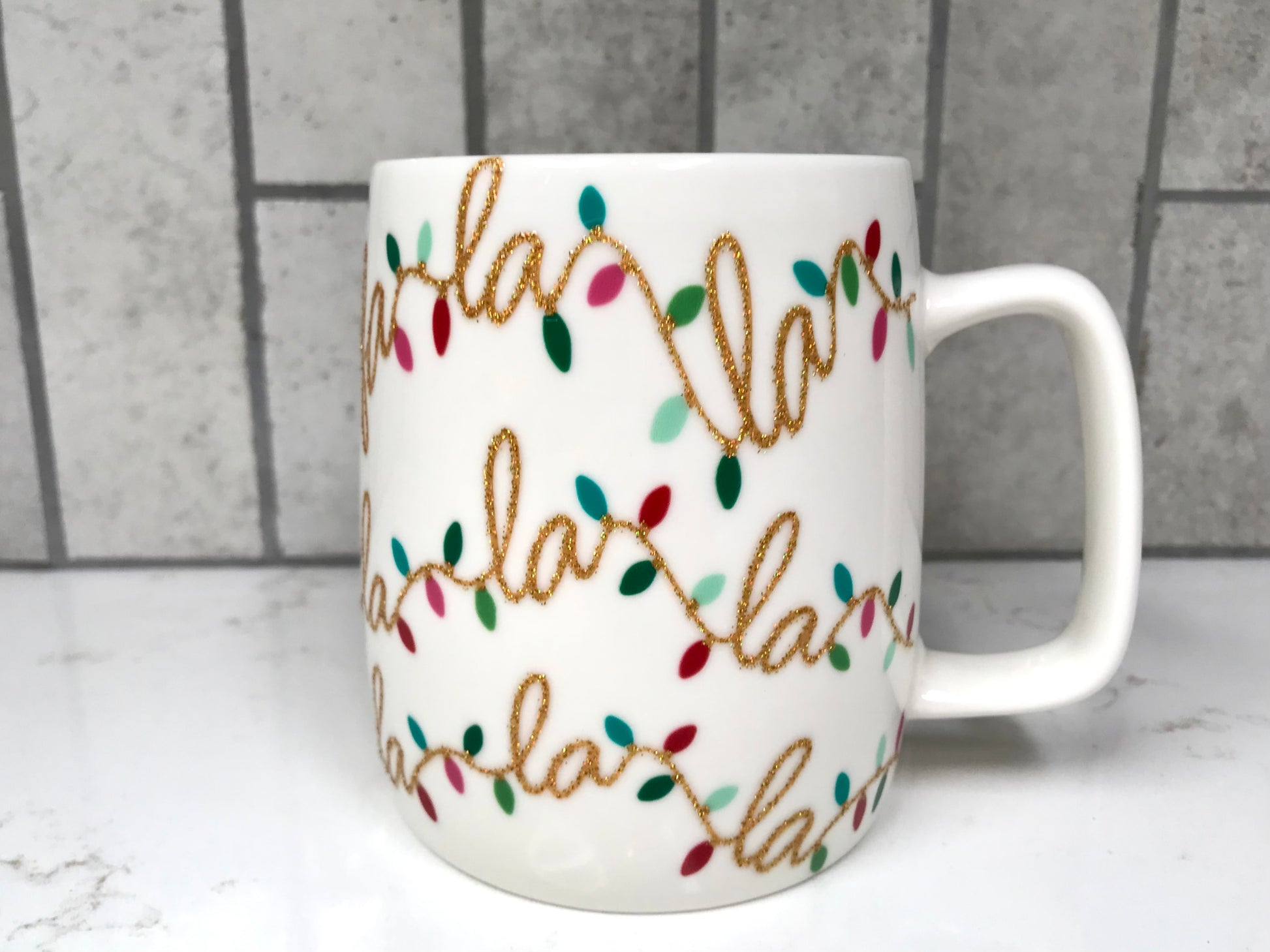 White mug with 'Fa La La La' text and colorful lights design on a marble surface with tiled wall background