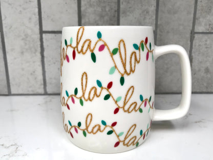 White mug with 'Fa La La La' text and colorful lights design on a marble surface with tiled wall background