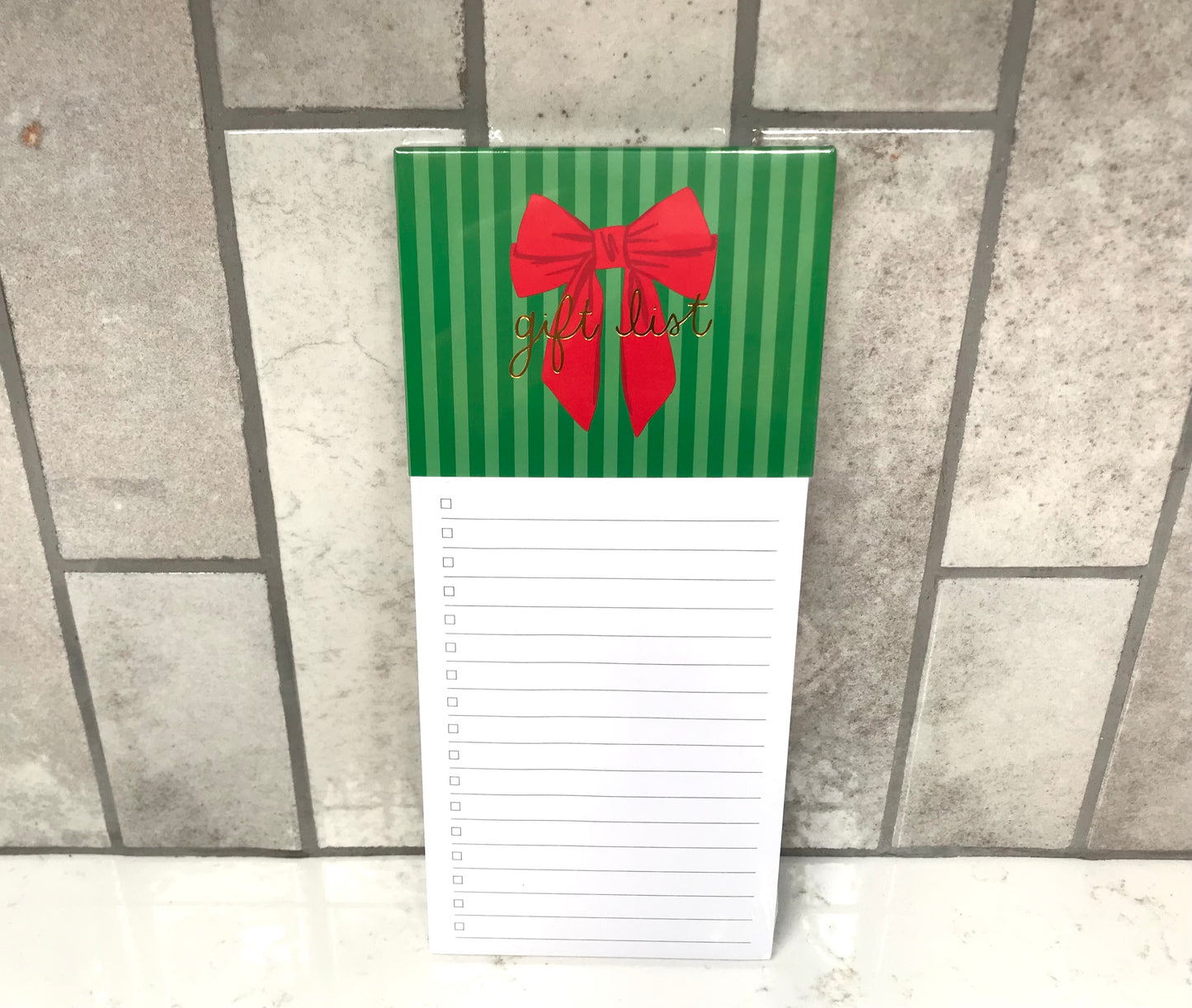 Notepad with green striped cover and red bow, featuring 'gift list' text, on a tiled floor.