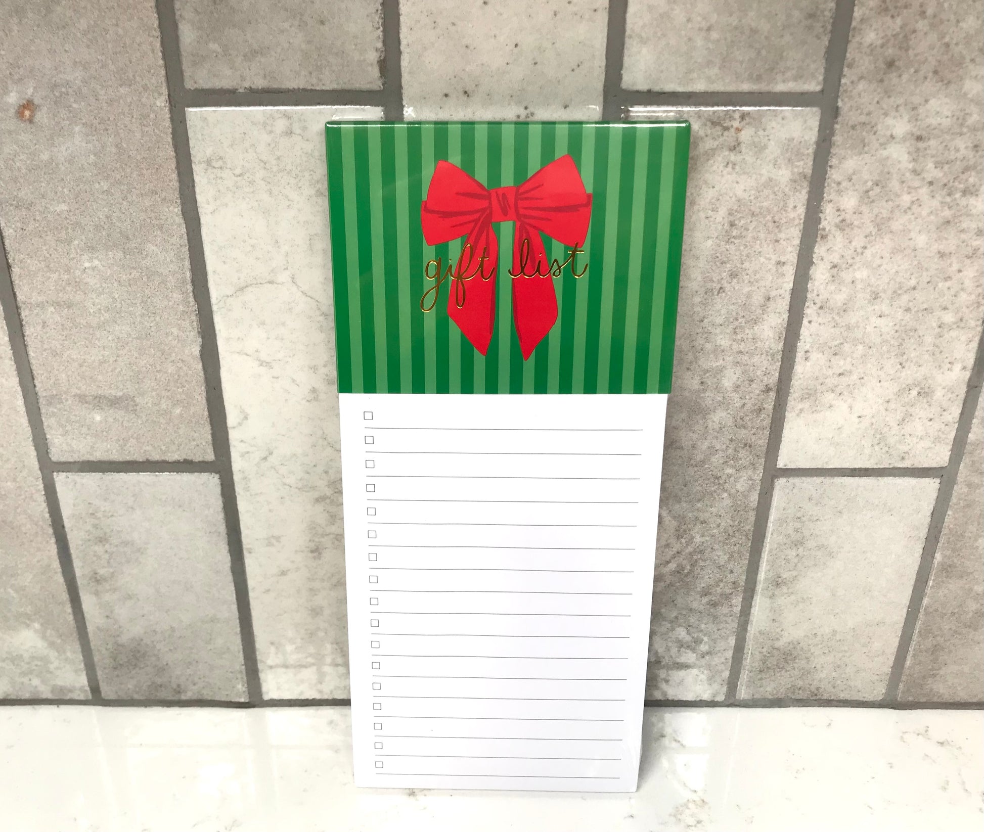 Notepad with green striped cover and red bow, featuring 'gift list' text, on a tiled floor.