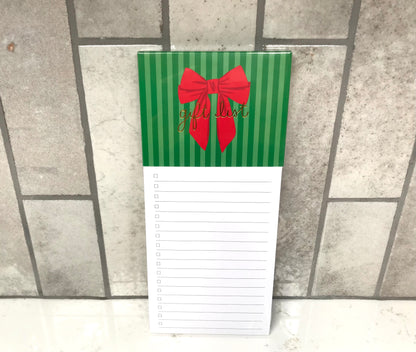 Notepad with green striped cover and red bow, featuring 'gift list' text, on a tiled floor.