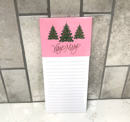 Notepad with Christmas tree design and 'Very Merry' text on a pink background, placed on a tiled surface.