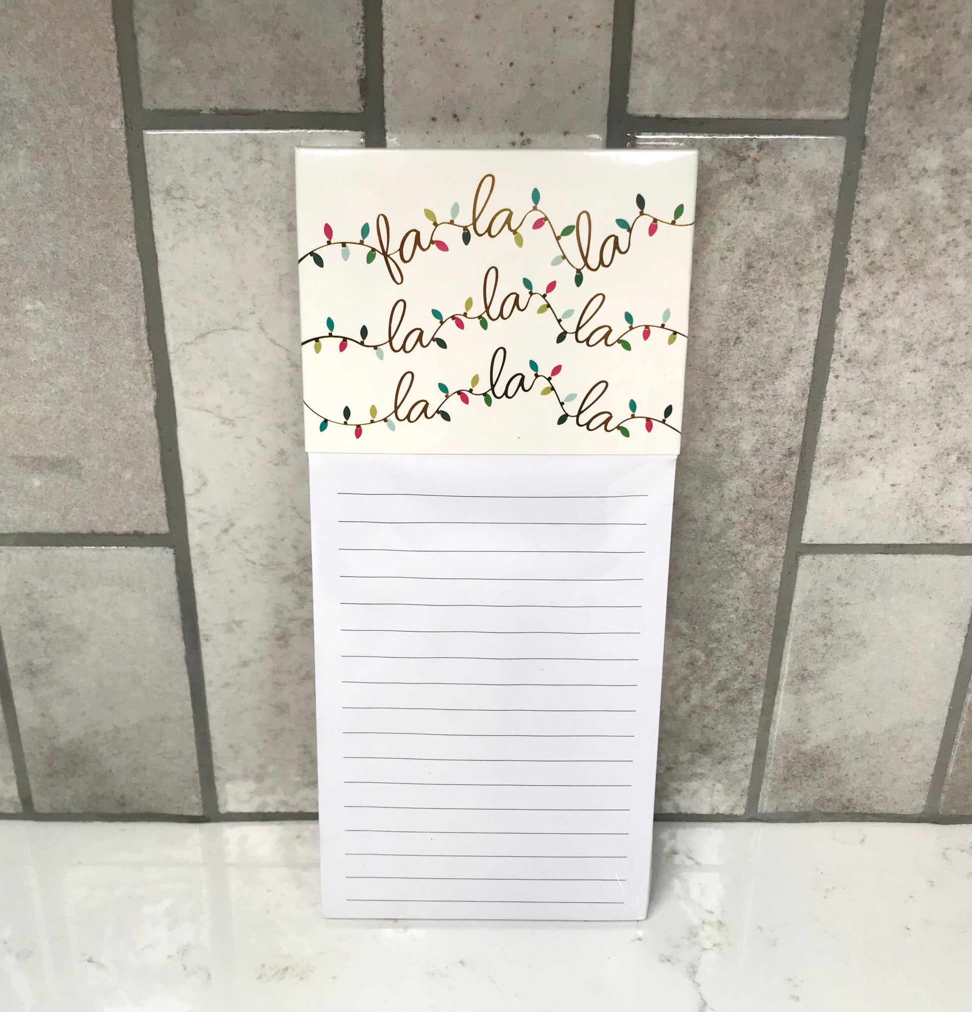 Notepad with 'fa la la la' text and Christmas lights design on a tiled floor background