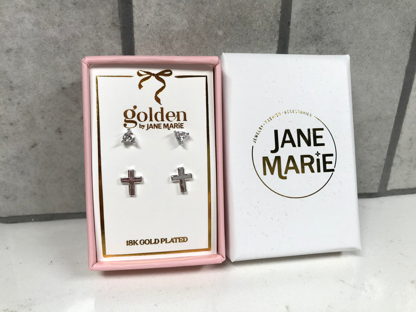 Kids 18K gold-plated earring set by Jane Marie shown in a pink gift box, including crystal bow studs on top and small silver cross studs on bottom. White Jane Marie packaging sleeve placed beside the box on a light tile backdrop.