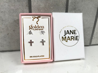 Kids 18K gold-plated earring set by Jane Marie shown in a pink gift box, including crystal bow studs on top and small silver cross studs on bottom. White Jane Marie packaging sleeve placed beside the box on a light tile backdrop.