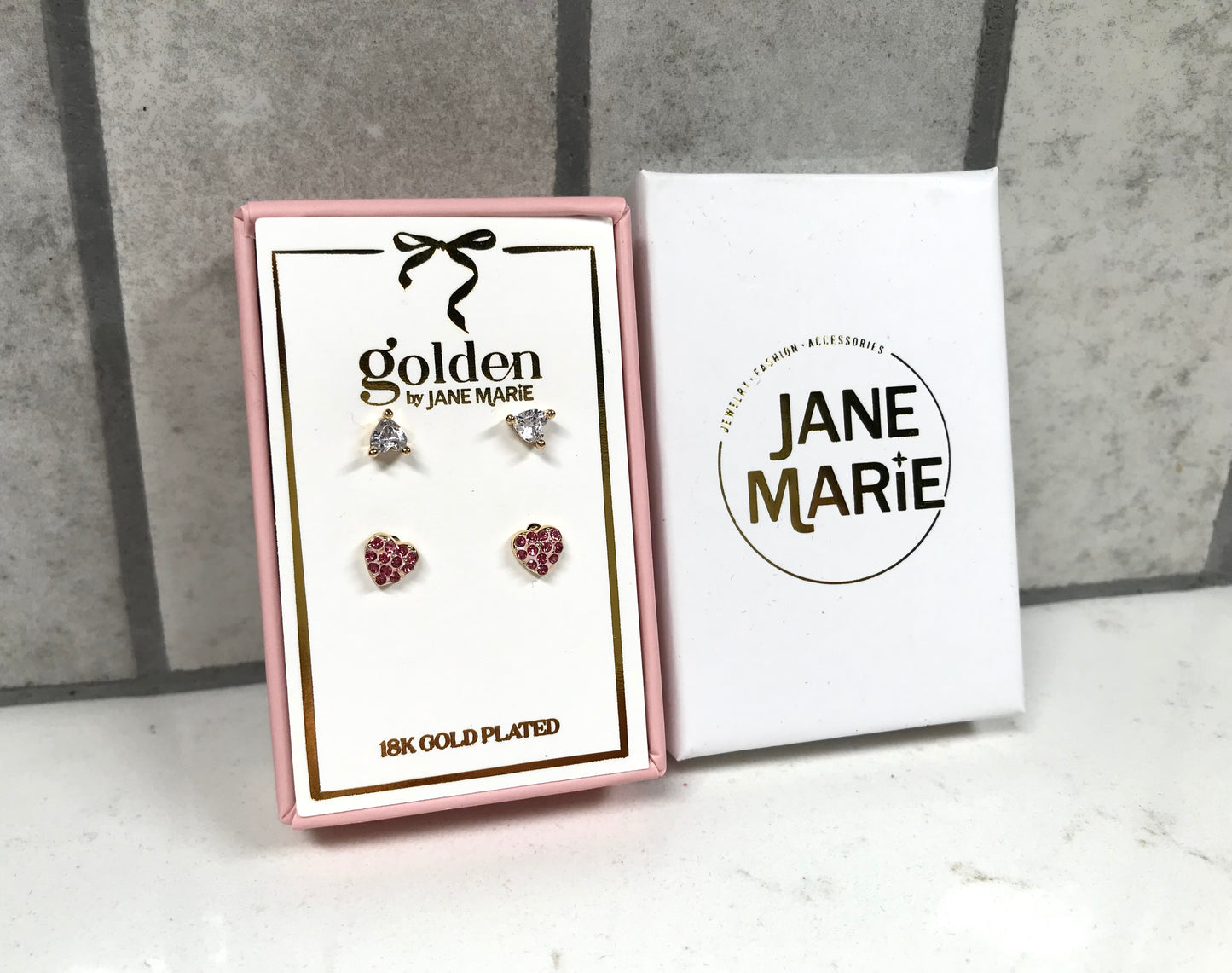 Kids 18K gold-plated earring set displayed in a pink Jane Marie gift box, featuring two pairs of studs: crystal bow studs on top and pink heart pavé studs on bottom. Box sits beside a white Jane Marie sleeve on a light tile background.