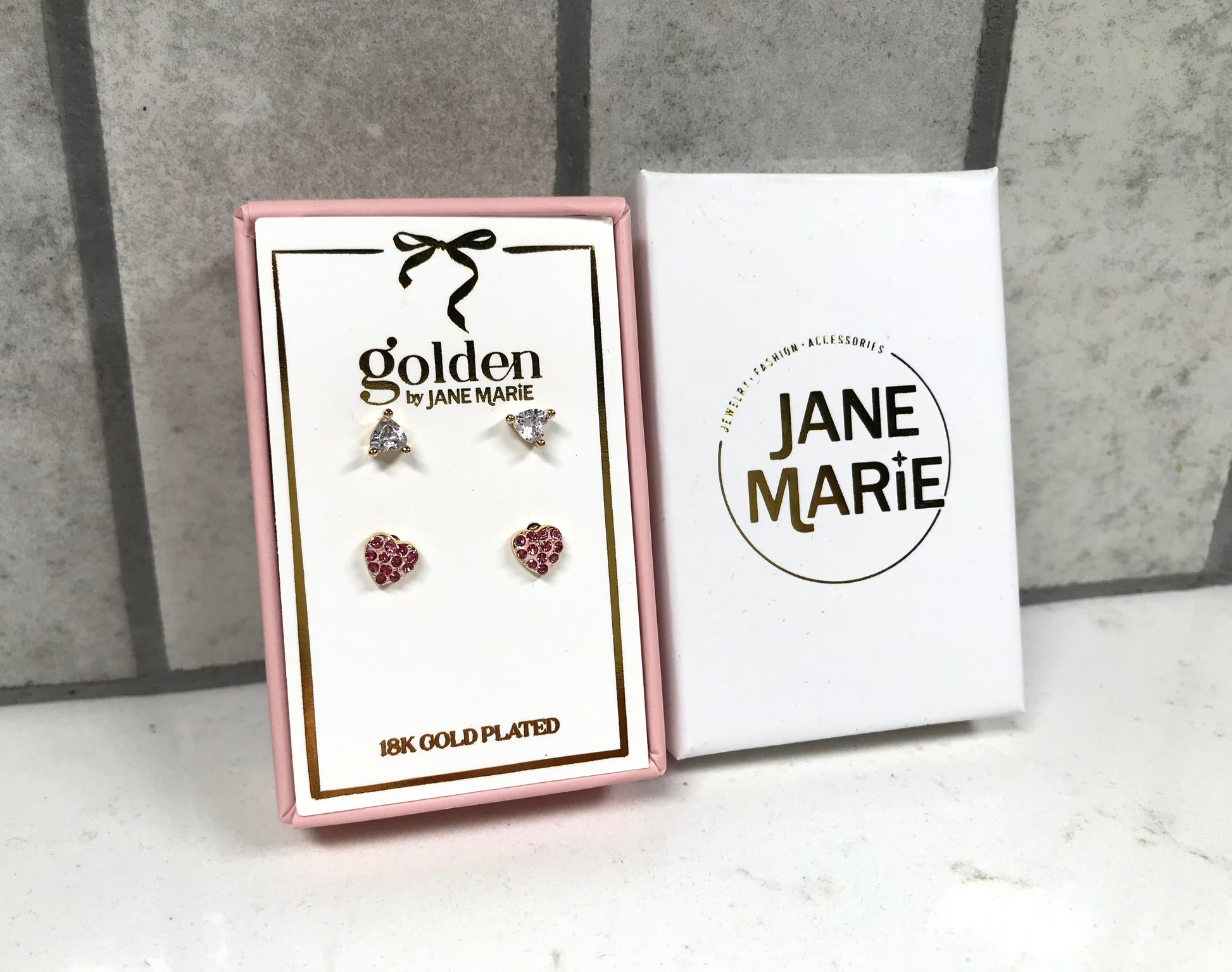 Kids 18K gold-plated earring set displayed in a pink Jane Marie gift box, featuring two pairs of studs: crystal bow studs on top and pink heart pavé studs on bottom. Box sits beside a white Jane Marie sleeve on a light tile background.