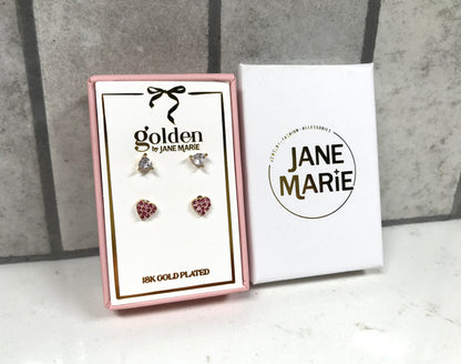 Kids 18K gold-plated earring set displayed in a pink Jane Marie gift box, featuring two pairs of studs: crystal bow studs on top and pink heart pavé studs on bottom. Box sits beside a white Jane Marie sleeve on a light tile background.
