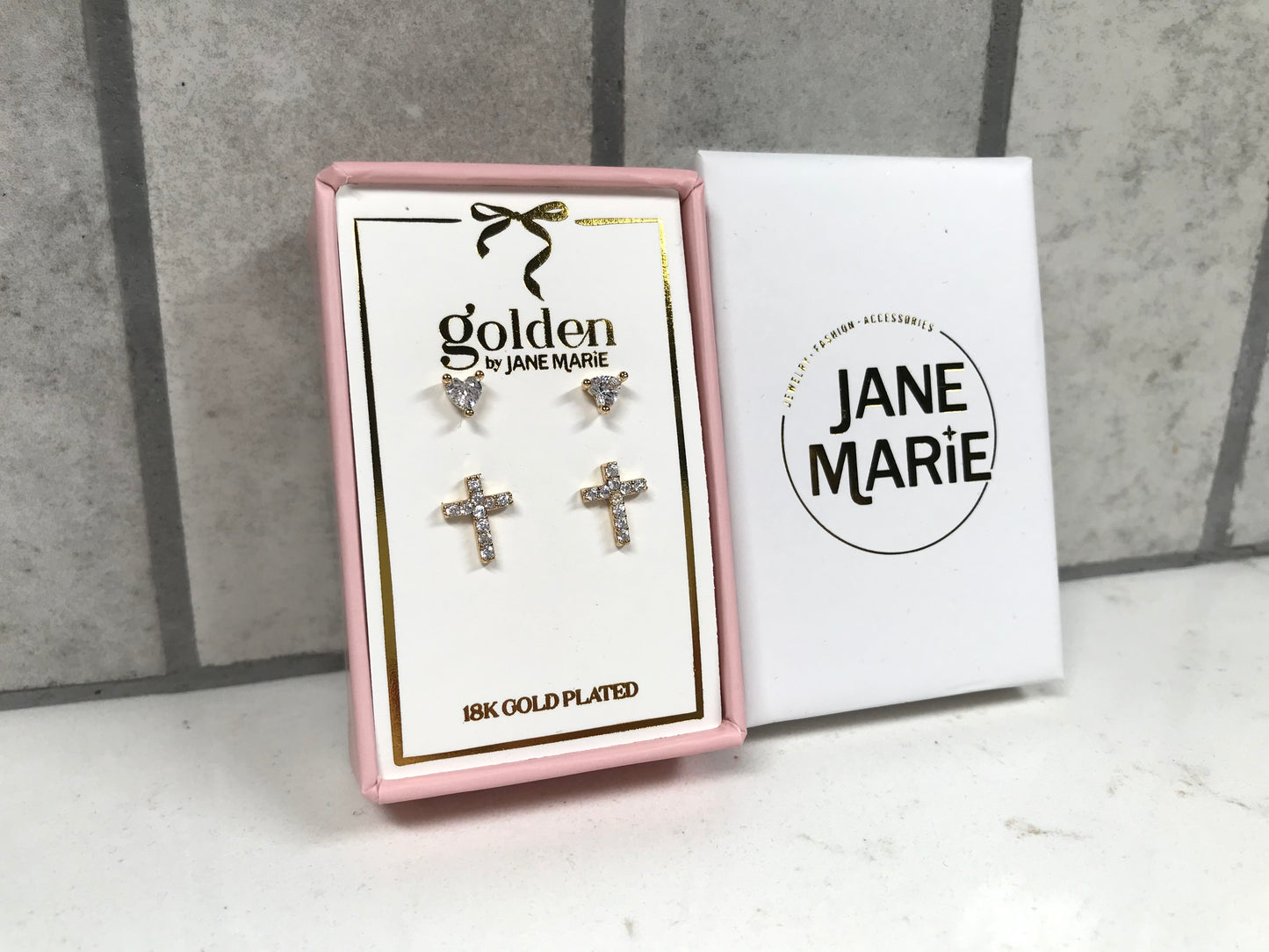 Kids 18K gold-plated earring set in a pink Jane Marie box featuring two pairs: crystal heart studs on top and gold pavé cross studs on the bottom. White Jane Marie sleeve displayed next to the box against a tile background.