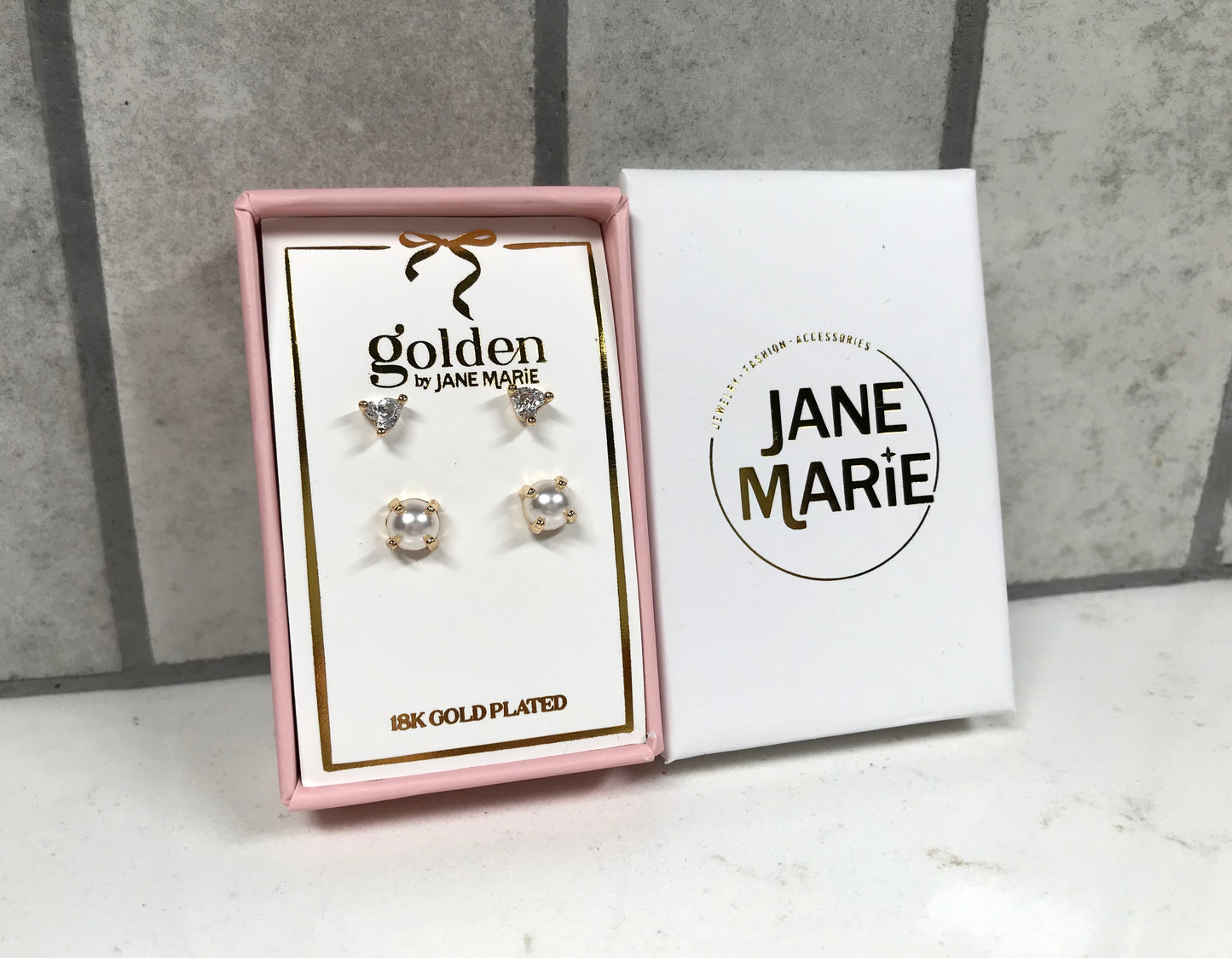 Kids 18K gold-plated earring set by Jane Marie in a pink box, including crystal heart studs on top and small pearl cluster studs on bottom. A white Jane Marie sleeve sits beside the box on a light tile background.