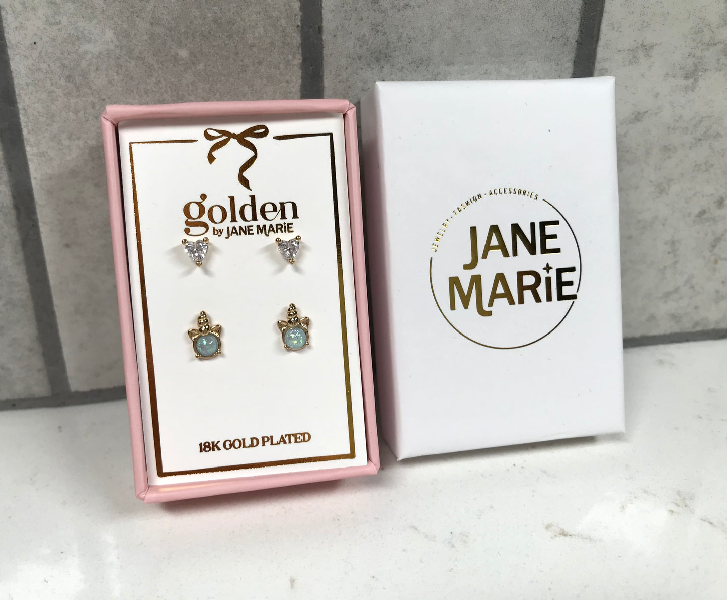Kids 18K gold-plated earring set by Jane Marie with crystal heart studs and blue turtle stud earrings, shown in a pink jewelry box beside white Jane Marie packaging.