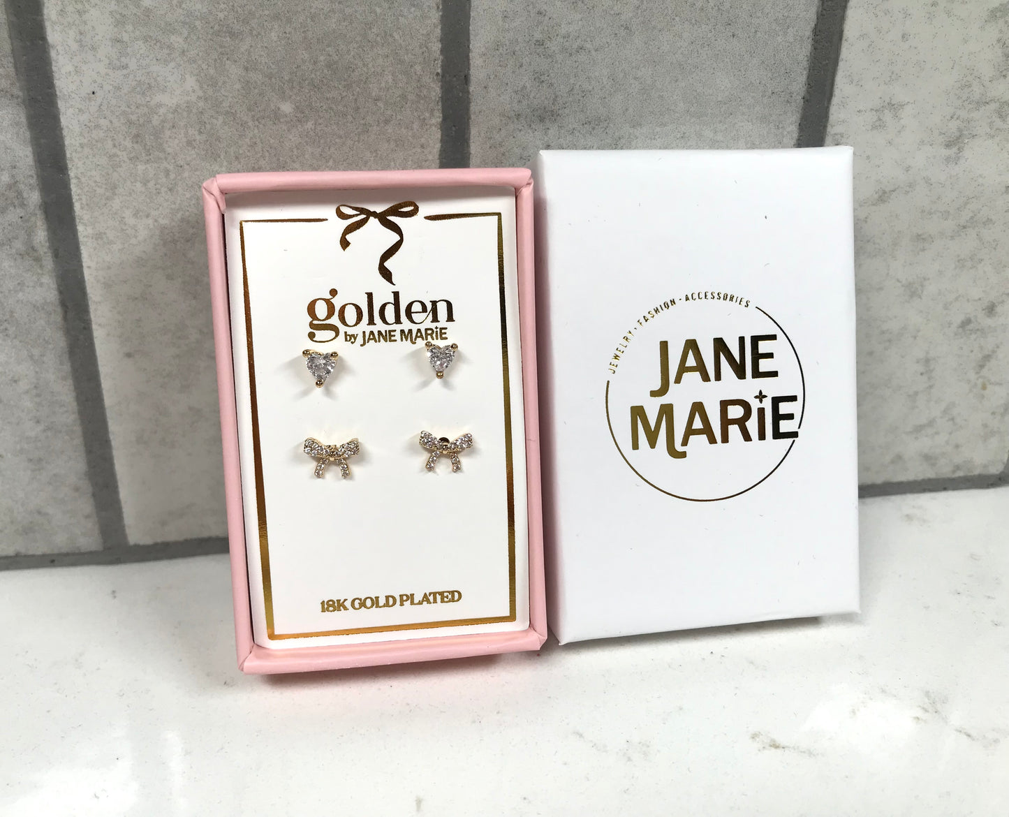 Kids 18K gold-plated earring set by Jane Marie including crystal heart studs and crystal bow studs, packaged in a pink gift box next to the white Jane Marie sleeve.