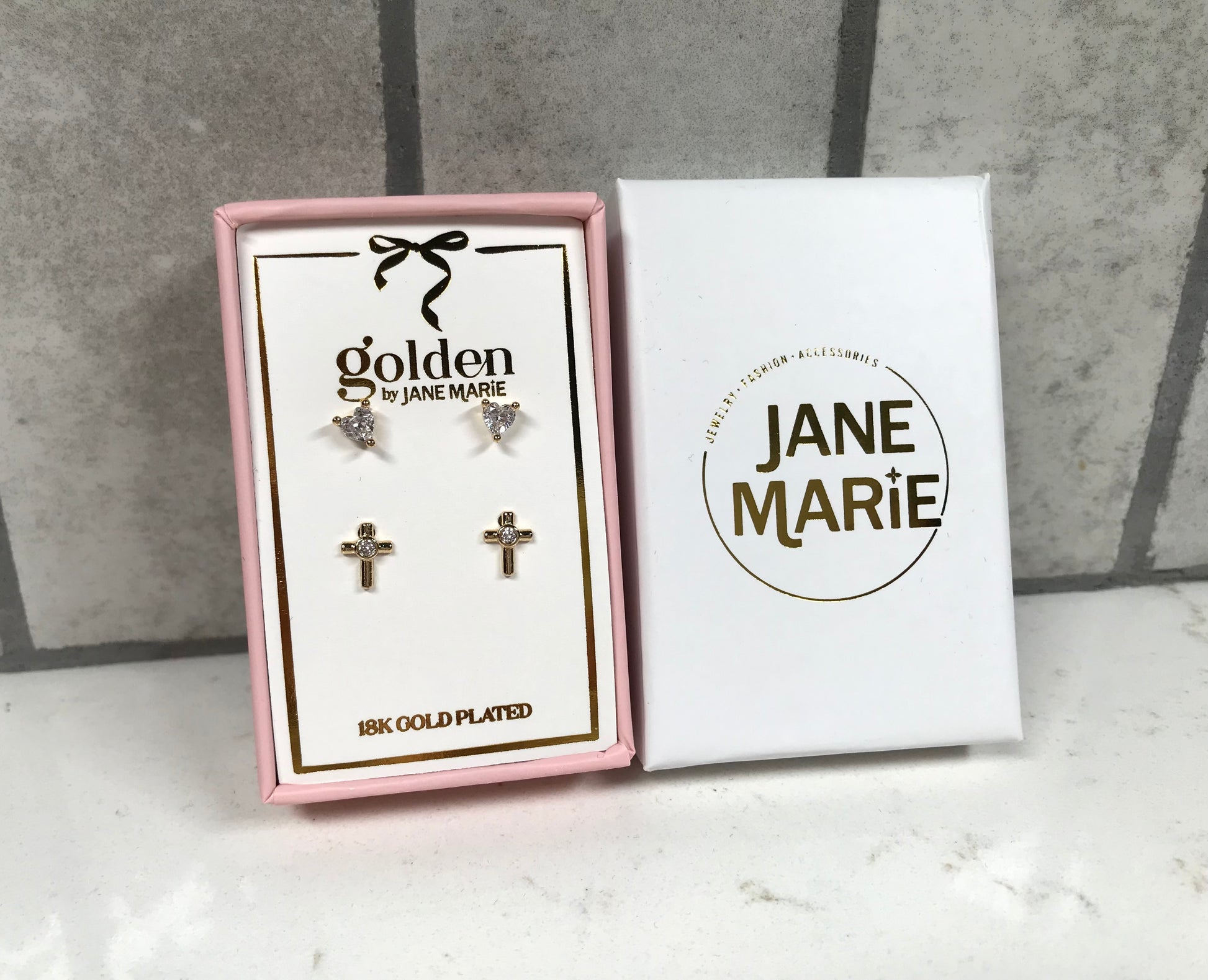 Kids 18K gold-plated earring set by Jane Marie featuring crystal heart studs and gold cross studs with crystal centers, displayed in a pink box with white Jane Marie sleeve.