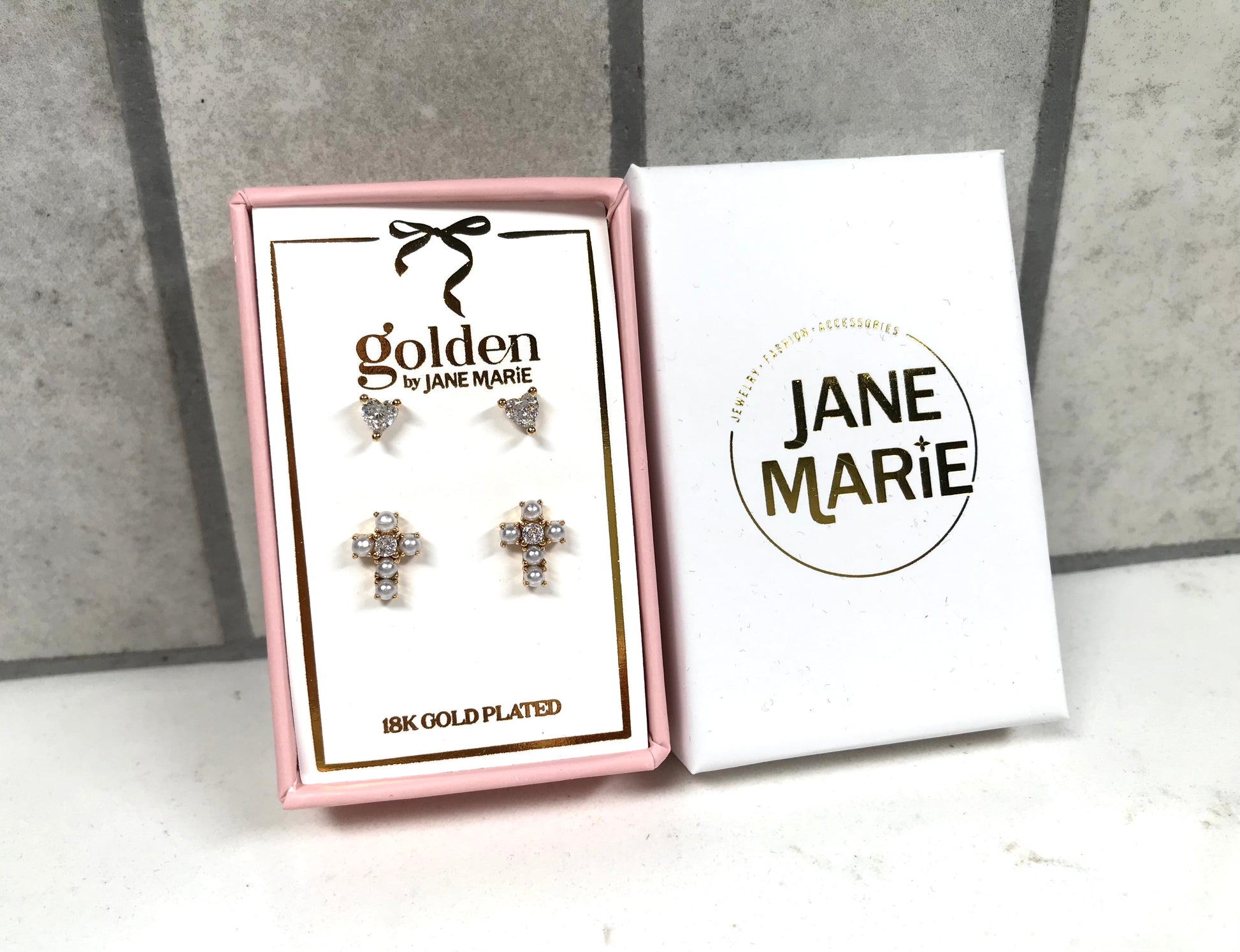 Kids 18K gold-plated stud earring set by Jane Marie, including crystal heart studs and clustered crystal cross studs, packaged in a pink gift box next to white Jane Marie branding sleeve.
