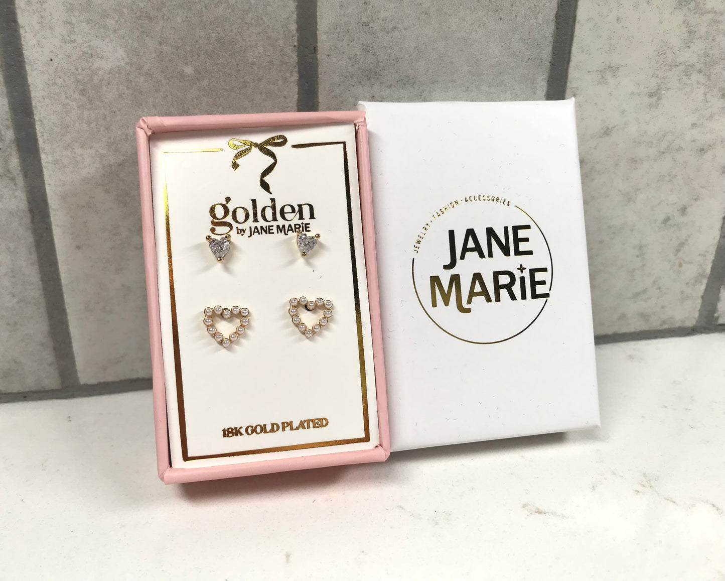 Kids 18K gold-plated earring set by Jane Marie featuring crystal heart studs and open heart pearl-accent studs, shown in a pink gift box beside the white Jane Marie sleeve.