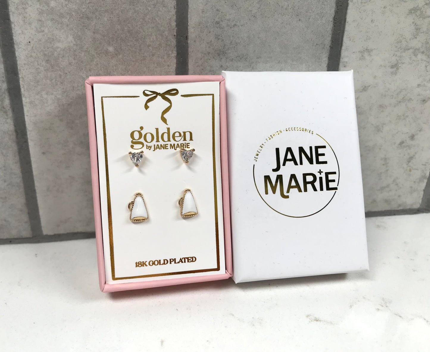 Kids 18K gold-plated earring set by Jane Marie with crystal heart studs and white megaphone studs, packaged in a pink jewelry box next to white Jane Marie sleeve.