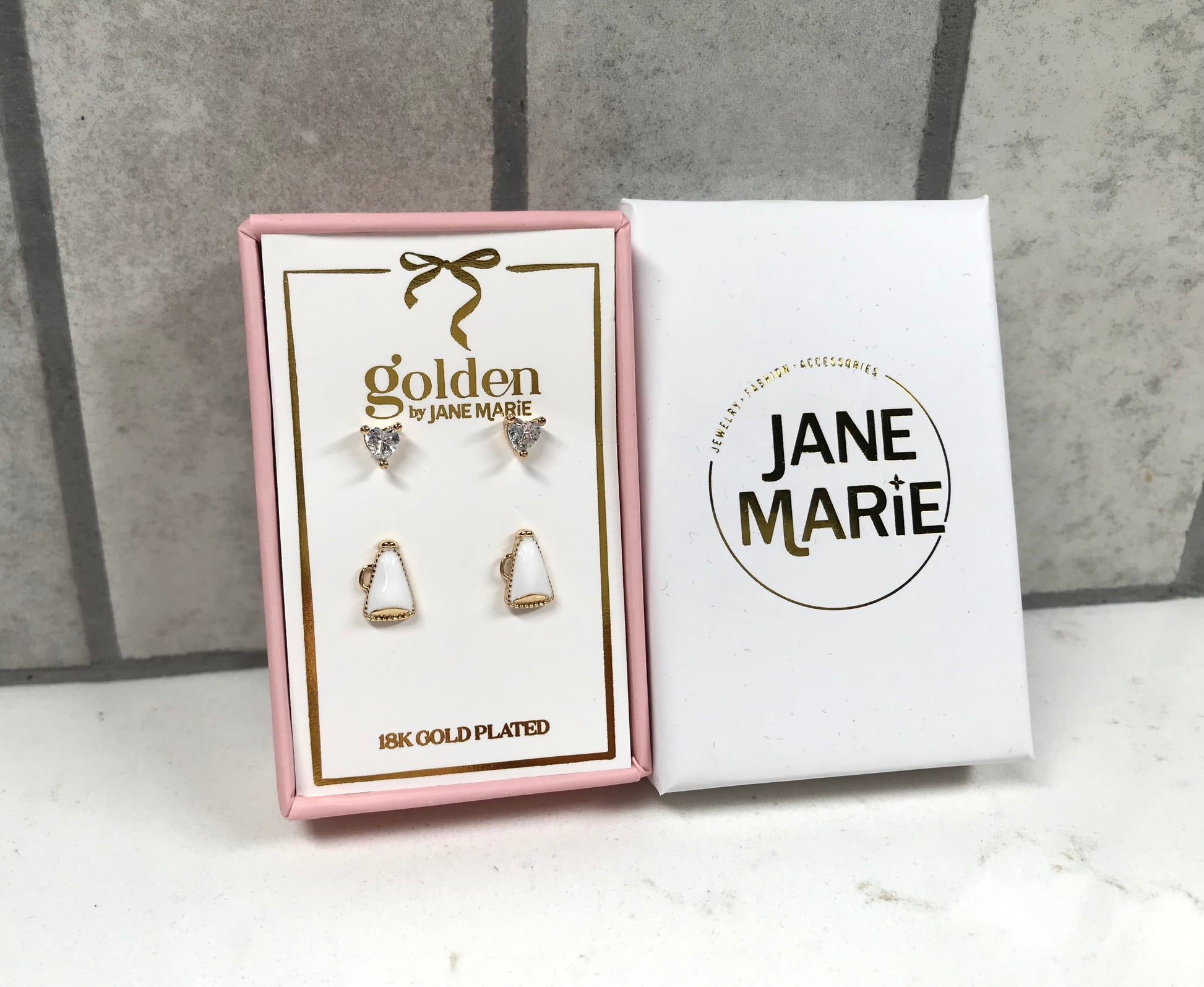 Kids 18K gold-plated earring set by Jane Marie with crystal heart studs and white megaphone studs, packaged in a pink jewelry box next to white Jane Marie sleeve.