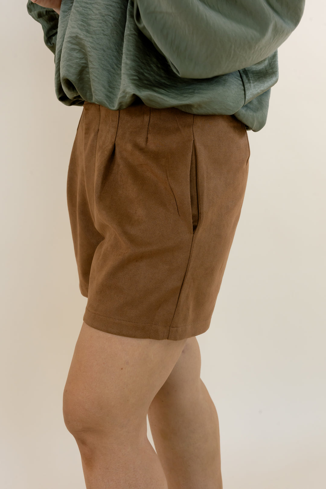 Camel brown faux suede pleated shorts for women featuring a tailored fit, front pleats, and a relaxed leg opening, styled with a sage green blouse.