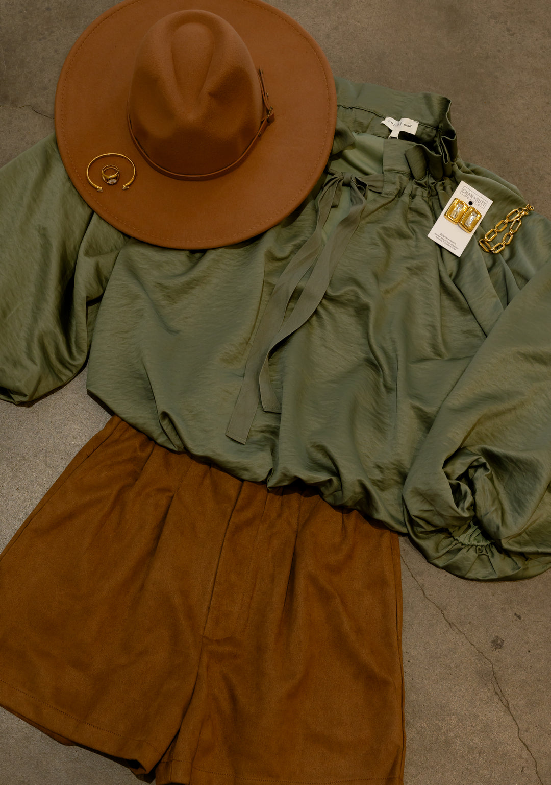 Camel brown faux suede pleated shorts styled with a sage green blouse, brown wide brim hat, and gold jewelry for a coordinated women&