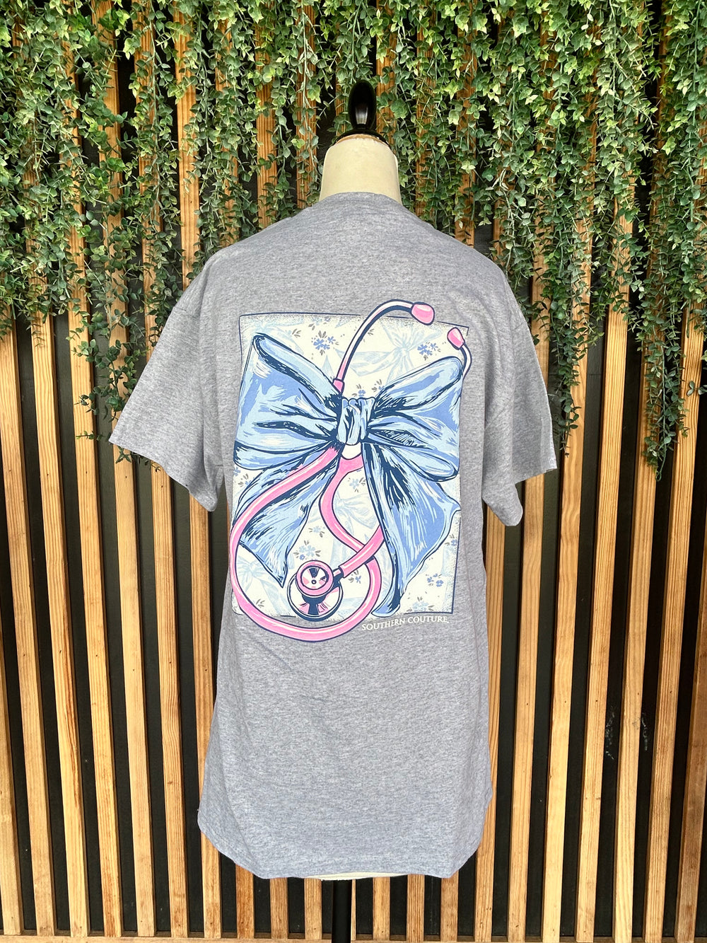 Back view of the Southern Couture Stethoscope Bow Graphic Tee displayed on a mannequin, showcasing a large blue bow and pink stethoscope graphic on a heather gray short sleeve t-shirt.