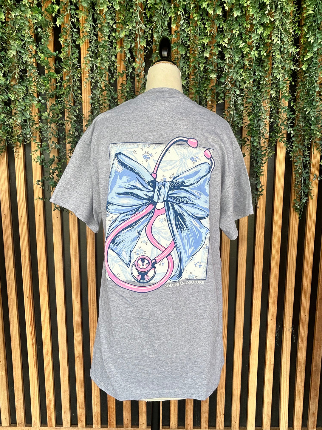 Back view of the Southern Couture Stethoscope Bow Graphic Tee displayed on a mannequin, showcasing a large blue bow and pink stethoscope graphic on a heather gray short sleeve t-shirt.