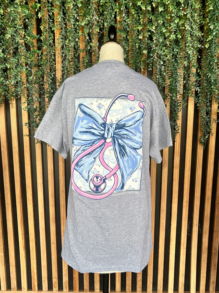Back view of the Southern Couture Stethoscope Bow Graphic Tee displayed on a mannequin, showcasing a large blue bow and pink stethoscope graphic on a heather gray short sleeve t-shirt.