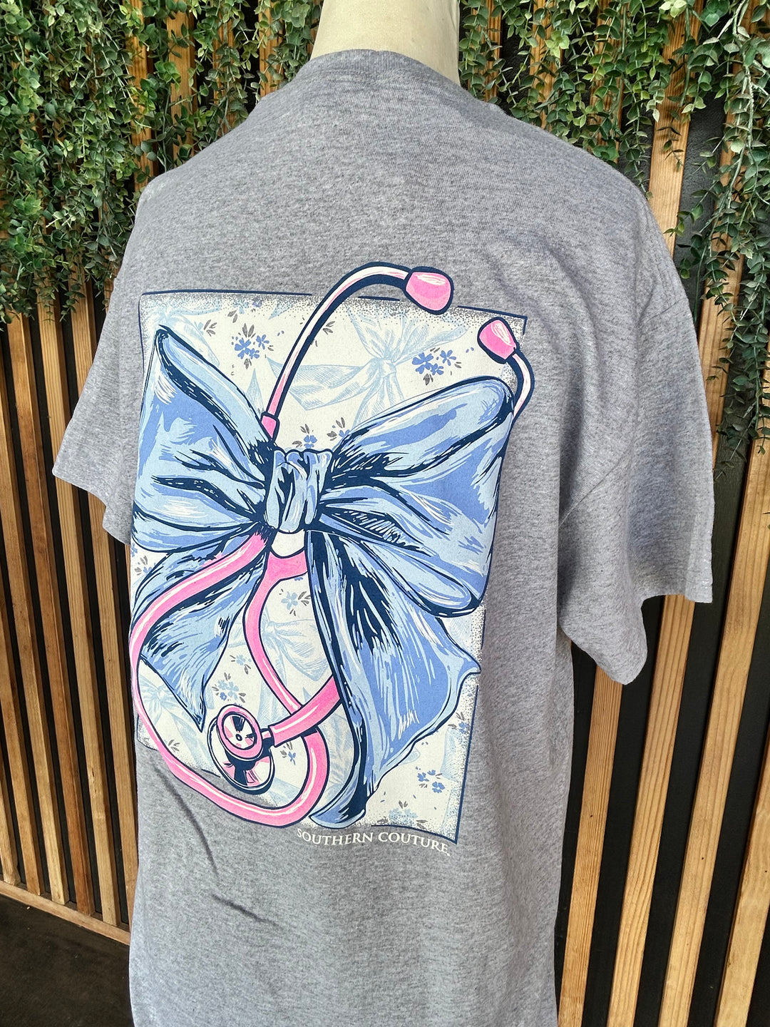 Back close-up of the Southern Couture Stethoscope Bow Graphic Tee featuring a large blue bow intertwined with a pink stethoscope on a heather gray 100% cotton t-shirt.