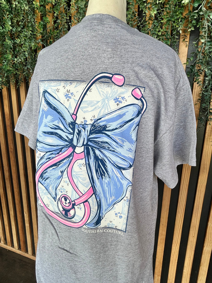 Back close-up of the Southern Couture Stethoscope Bow Graphic Tee featuring a large blue bow intertwined with a pink stethoscope on a heather gray 100% cotton t-shirt.