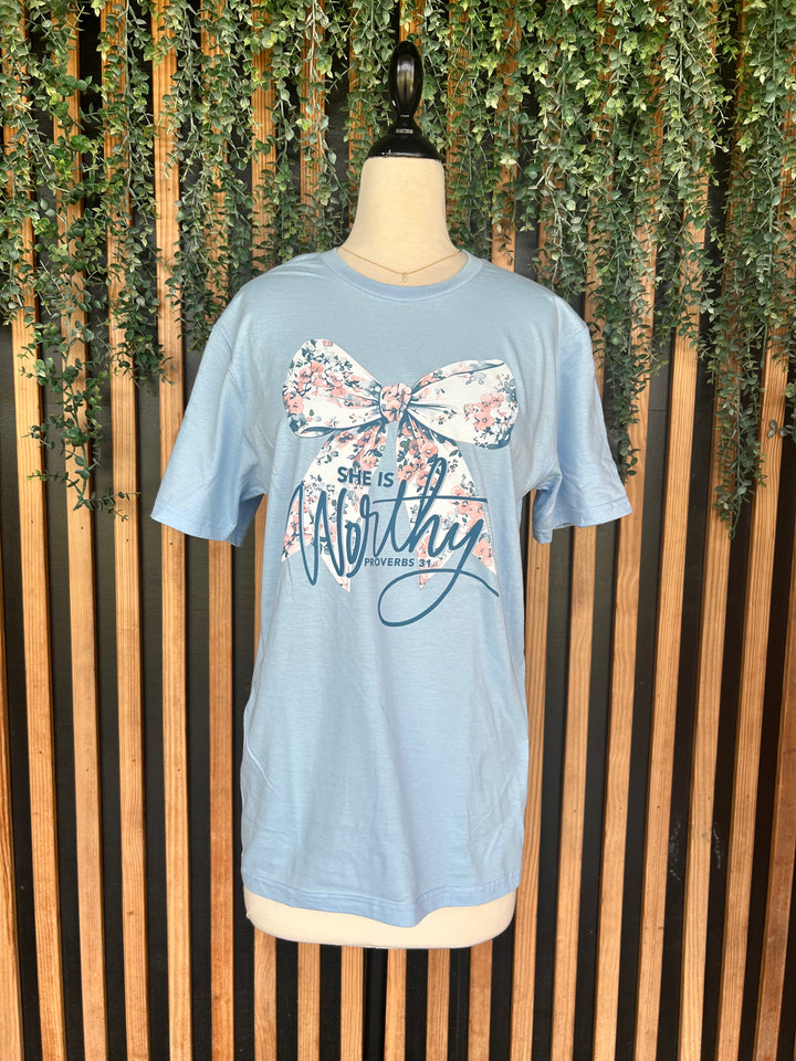 Light blue She Is Worthy Floral Bow Graphic Tee displayed on a mannequin against a wood slat and greenery backdrop. This 100% cotton Christian graphic t-shirt features a large floral bow design, Proverbs 31:25 message, short sleeves, and a classic crew neckline.